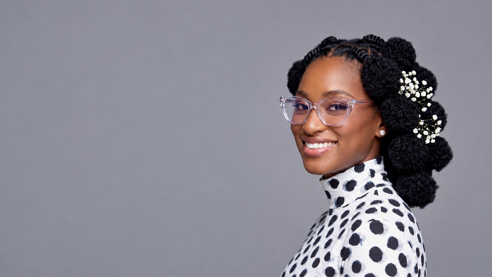 Portrait showing shoulders and upper chest, in clear glasses and a polka-dot blouse against a gray studio background